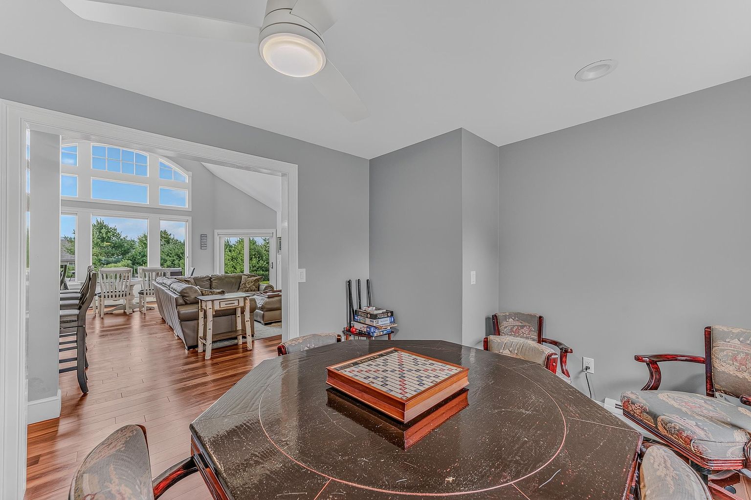 This dining room features a dark, circular table set for a game, surrounded by patterned upholstered chairs. The room is painted in a neutral gray tone and opens into a bright, sun-drenched living area with large windows and hardwood flooring. The perspective is from the dining area looking through the doorway into the adjacent living space, creating an open and inviting atmosphere.