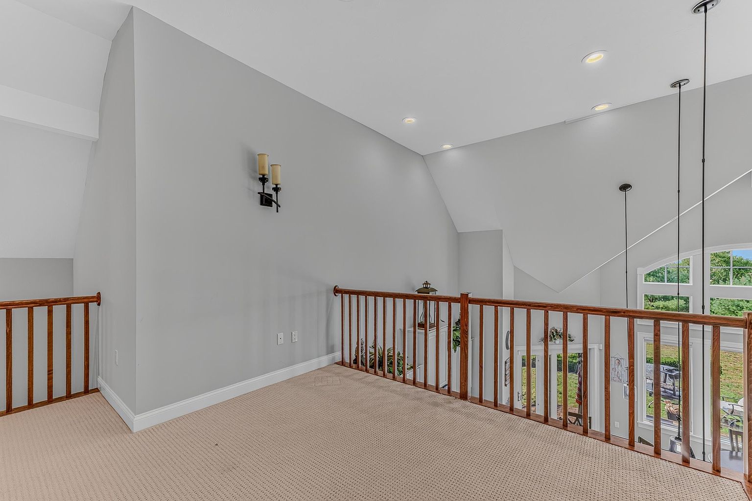 This image captures a carpeted loft area overlooking a lower level, defined by a wooden railing and high, vaulted ceilings. The space is painted in a neutral light gray, accented by a wall-mounted light fixture and recessed lighting. The perspective provides a clear view of the open floor plan below, emphasizing the airy, spacious feel of the home's architecture.