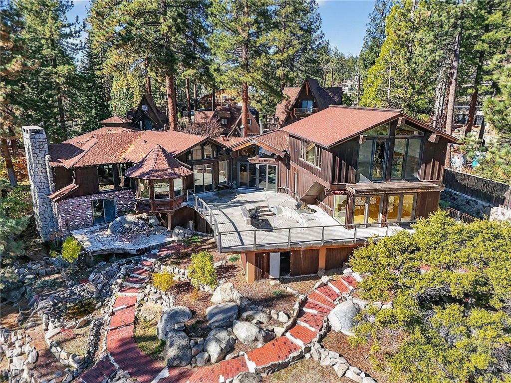 This aerial view showcases a multi-level home nestled among tall trees. The property features a combination of wood and brick exterior, a spacious deck, and a landscaped yard with stone pathways and mature greenery. The overall impression is one of a secluded and luxurious mountain retreat.