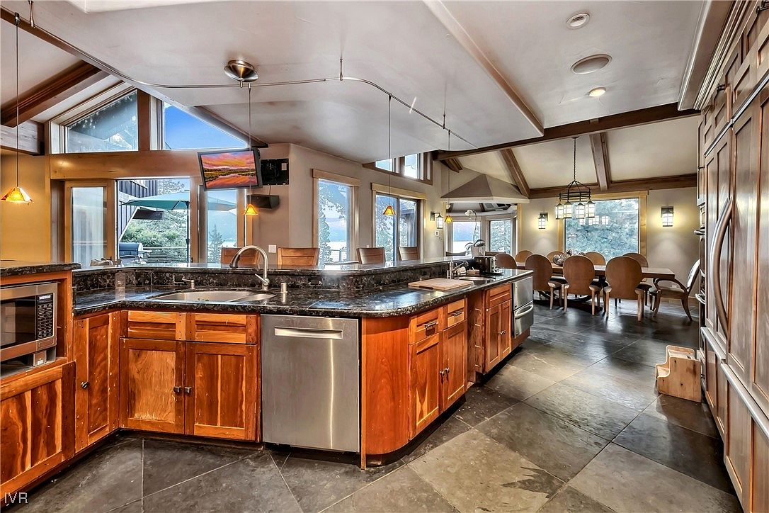 This interior shot showcases a spacious kitchen with warm wood cabinetry, dark granite countertops, and stainless steel appliances. The kitchen island provides ample counter space, and large windows offer scenic views. The adjacent dining area is visible, creating an open and inviting atmosphere.