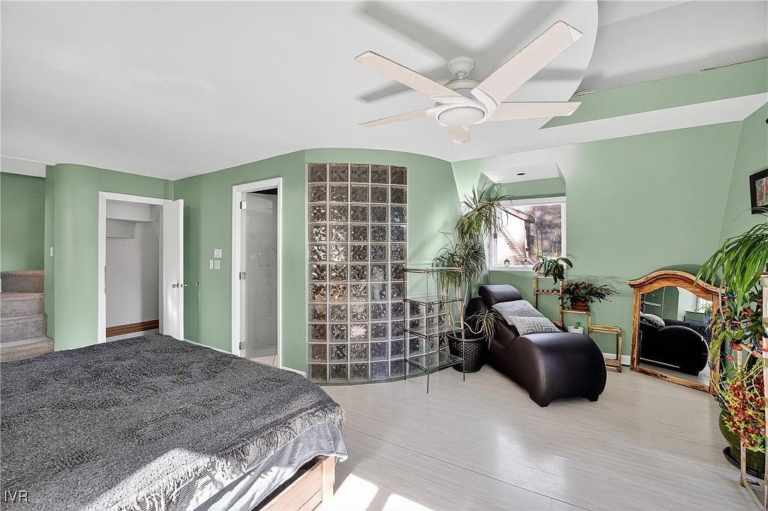 This is a primary bedroom featuring a unique architectural design with curved walls and a glass block partition. The room is painted in a soft green hue, complemented by light-colored wood flooring. A chaise lounge sits near a window, and a ceiling fan provides air circulation, creating a serene and inviting atmosphere.