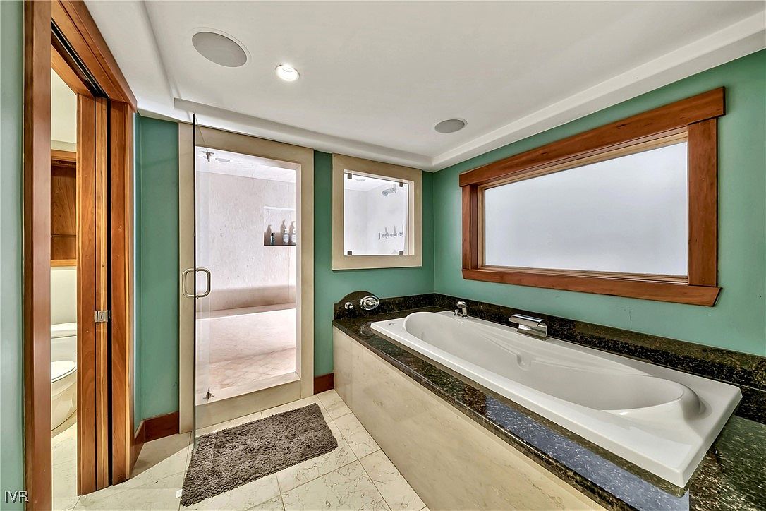 This is a primary bathroom featuring a large soaking tub with a dark granite countertop and a wood-framed window above. A glass-enclosed shower is visible through a doorway, and the walls are painted in a calming green hue. The overall impression is luxurious and spa-like.