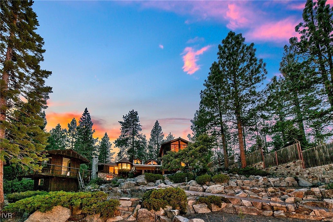 This image showcases the front of a charming mountain home at dusk, nestled among tall pine trees and a rock garden. The house features warm lighting emanating from its windows, creating an inviting atmosphere. The landscape is artfully arranged with rocks and greenery, enhancing the property's natural appeal.