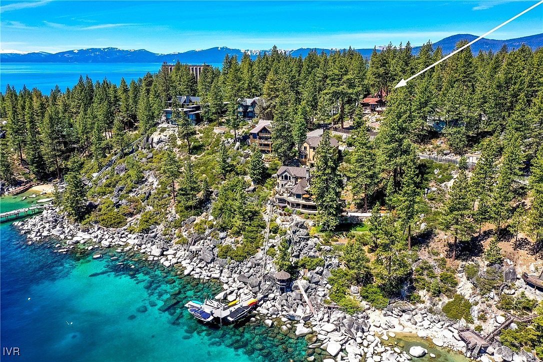 This aerial shot showcases a stunning lakeside property with multiple dwellings nestled among lush trees. The turquoise water contrasts beautifully with the rocky shoreline and the verdant landscape. A private dock with boats adds to the property's appeal, suggesting a luxurious and secluded retreat.