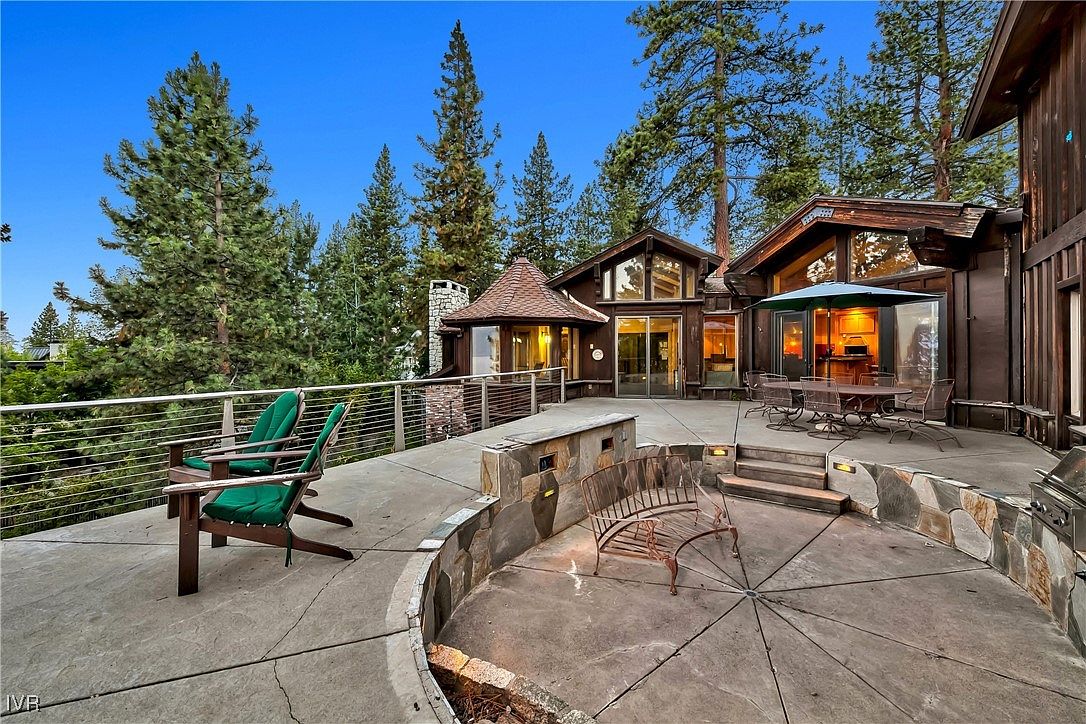This image showcases a spacious outdoor patio and deck area of a luxurious home. The patio features a unique fire pit area with a curved stone bench, along with a dining set under an umbrella, perfect for entertaining. The deck offers additional seating with comfortable chairs, all surrounded by lush trees, creating a serene and inviting outdoor living space.