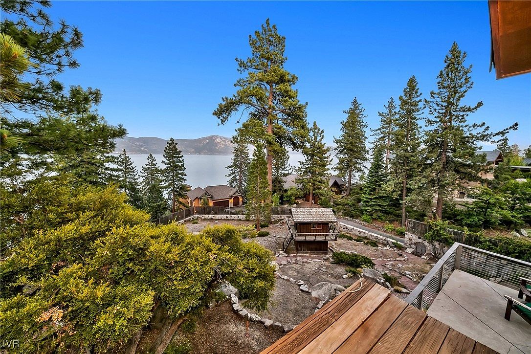 This image showcases a beautifully landscaped yard with mature trees, shrubs, and a charming wooden structure, possibly a playhouse or gazebo. The yard features natural rock formations and a winding pathway, leading to a view of a serene lake and distant mountains. A wooden deck provides an elevated vantage point, enhancing the property's appeal.