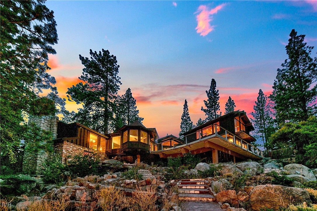 This image showcases the front exterior of a unique, multi-level home nestled among trees and rock formations. The architecture features a blend of wood and stone, with large windows offering views from various rooms. A stone and wooden staircase leads up to the house, blending seamlessly with the natural landscape, creating a sense of secluded luxury.