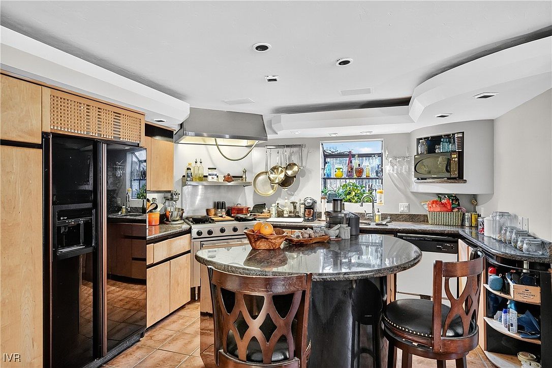 This is a well-appointed kitchen featuring light wood cabinetry, stainless steel appliances, and a unique circular island with a dark countertop and decorative seating. The kitchen is well-lit, with a window providing natural light and a view to the outside. The overall impression is one of functionality and style.