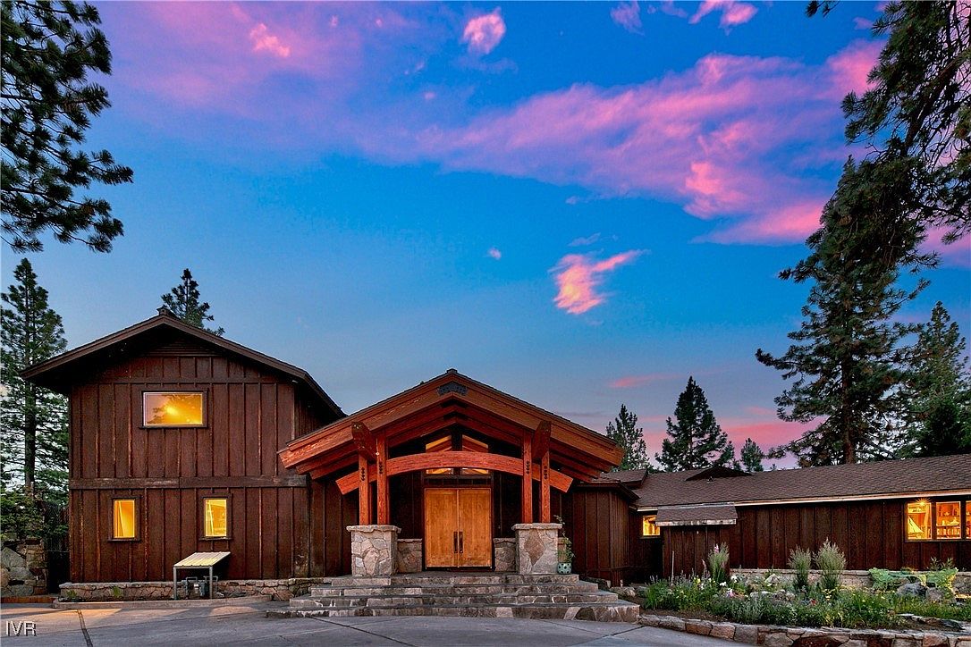 This is a front exterior view of a large, rustic-style home with a prominent wooden entryway and stone accents. The house features dark wood siding and a multi-level design, set against a backdrop of trees and a colorful sky at dusk. The overall impression is one of a well-maintained and inviting property.