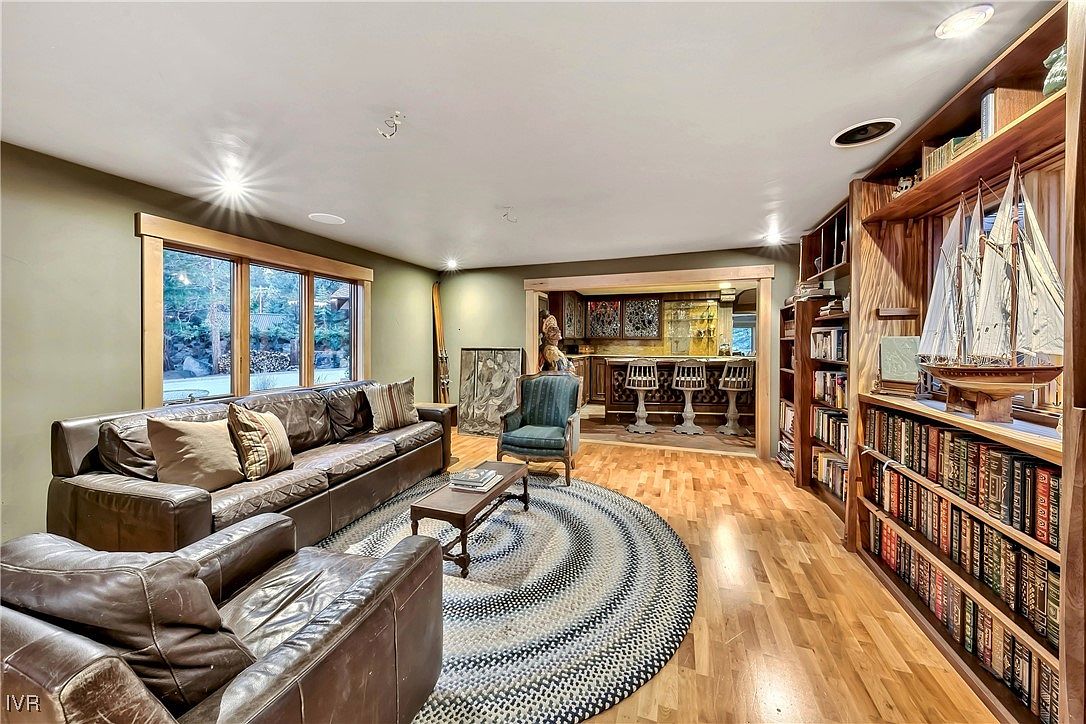 This is an interior shot of a living room featuring a brown leather sofa and armchair set around a round braided rug. A wooden coffee table sits in the center, and a built-in bookcase with a model sailboat adds character. In the background, there's a bar area with stools, creating a cozy and inviting atmosphere.