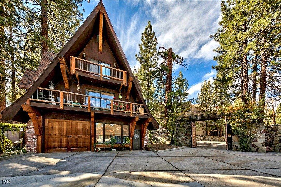 This is a front view of a charming A-frame house with a rustic, cabin-like aesthetic. The house features multiple balconies with wooden railings, a large wooden garage door, and a stone-paved driveway. The surrounding landscape includes mature trees and a stone archway, adding to the property's appeal and sense of privacy.
