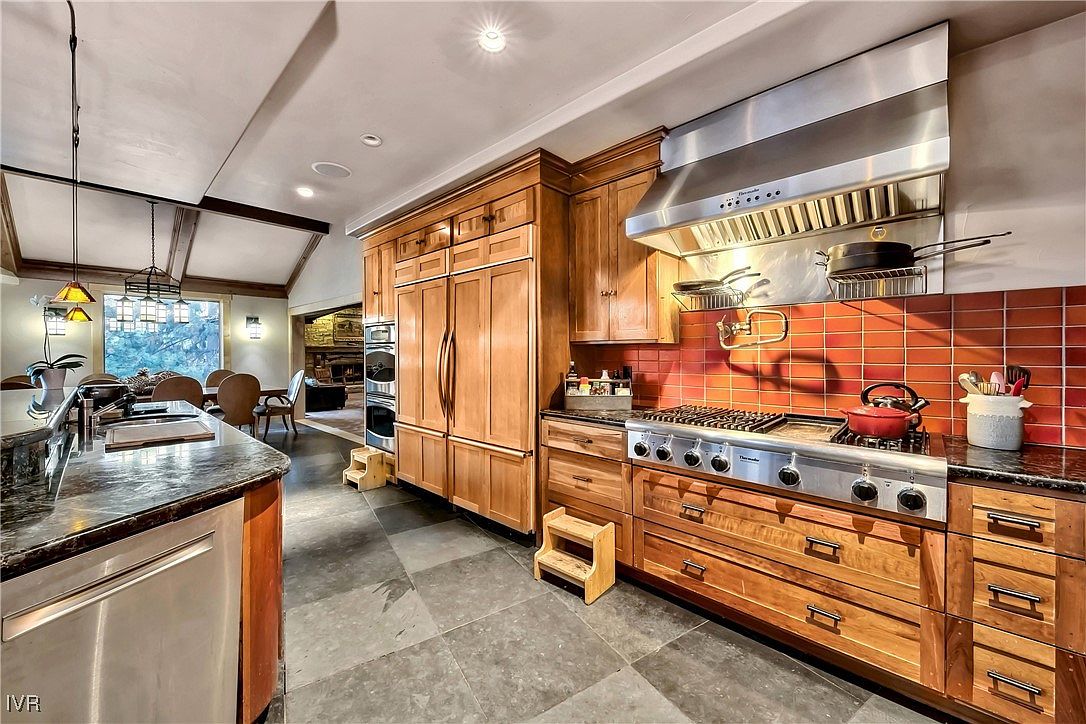 This is a well-appointed kitchen featuring custom wood cabinetry, stainless steel appliances, and granite countertops. The kitchen includes a large range with a stainless steel hood, a built-in refrigerator, and double ovens. The flooring is a dark stone tile, and there are two small wooden step stools to assist with reaching higher cabinets. The perspective is from the side of the kitchen, showing the island and dining area in the background.