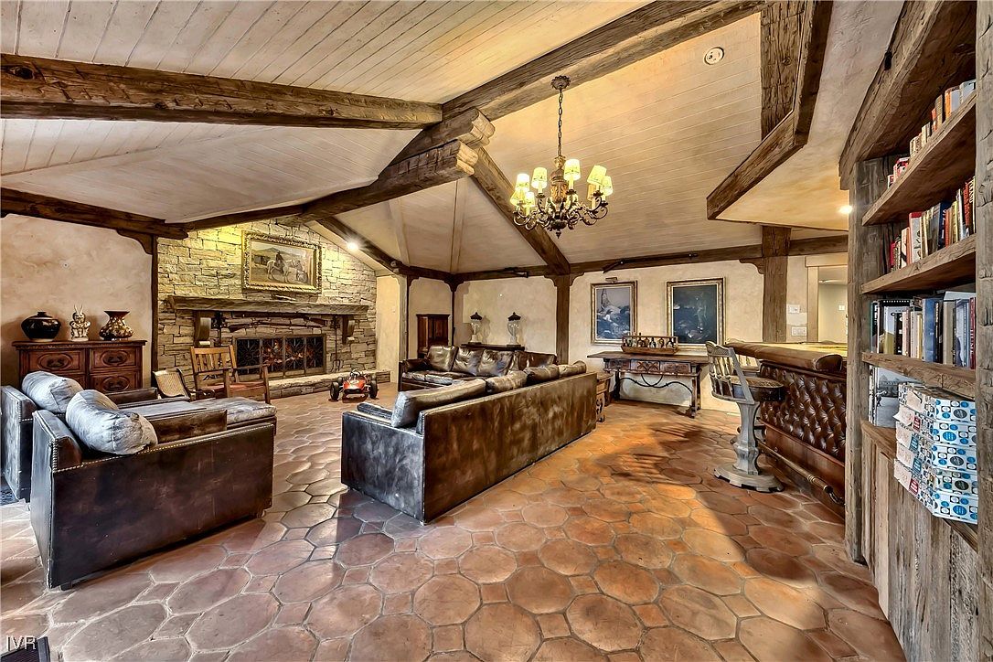 This is a spacious living room featuring exposed wooden beams, a stone fireplace, and a mix of leather sofas and chairs. The room is illuminated by a chandelier and natural light, creating a warm and inviting atmosphere. The flooring is a unique tile pattern, and there's a built-in bookshelf and bar area, adding to the room's character and functionality.