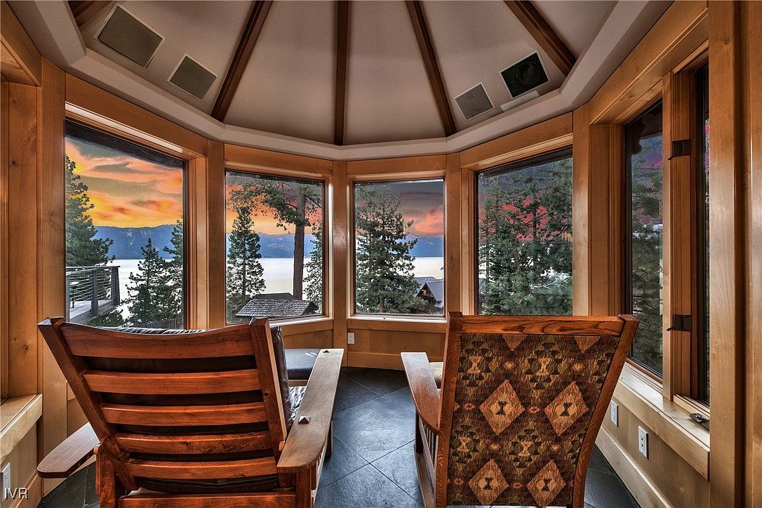 This sunroom features a panoramic view of a lake and mountains through large windows, creating a serene and inviting atmosphere. Two wooden chairs are positioned to take advantage of the view, enhancing the room's appeal as a relaxation space. The room's design incorporates natural wood finishes and a unique ceiling structure, adding to its architectural interest.
