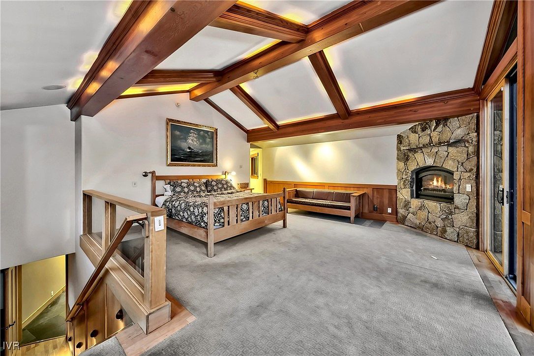 This primary bedroom features a vaulted ceiling with exposed wooden beams and recessed lighting, creating a warm and inviting atmosphere. A stone fireplace adds a rustic touch, while a large bed with patterned bedding anchors the space. The room also includes a seating area and access to an outdoor space through sliding glass doors, offering a luxurious and comfortable retreat.
