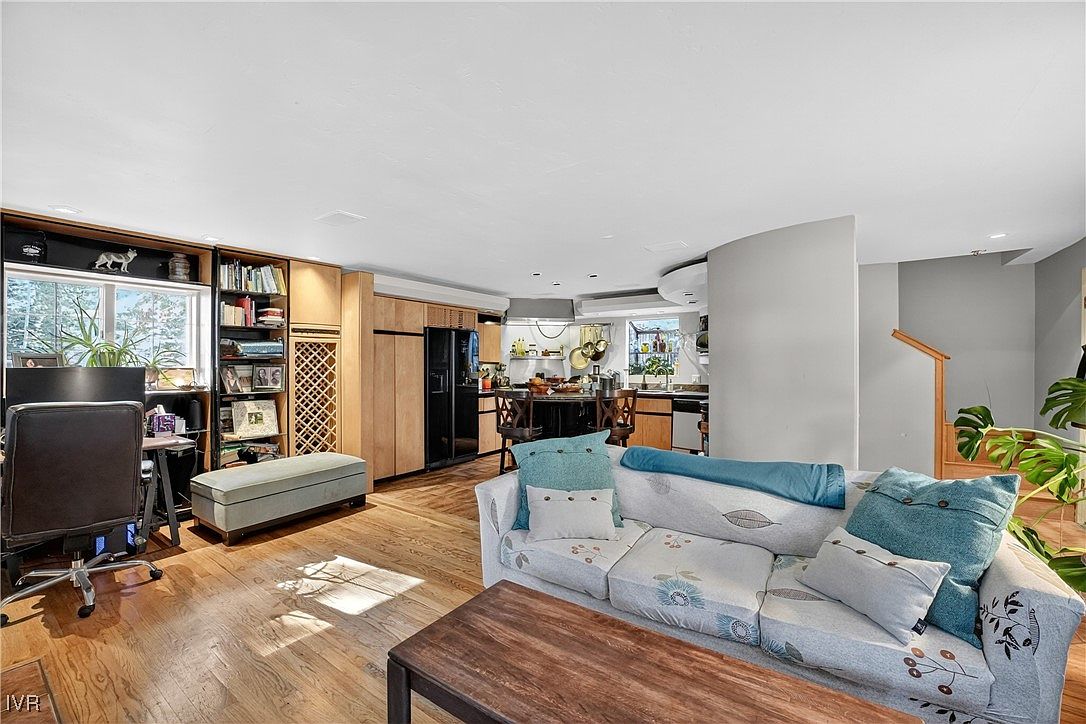 This is an interior shot of a living room that flows into a kitchen area. The living room features hardwood floors, a floral patterned sofa with throw pillows, and a dark wood coffee table. The kitchen area has wooden cabinets and black appliances, with a dining table visible in the background. The overall impression is a cozy and inviting living space.