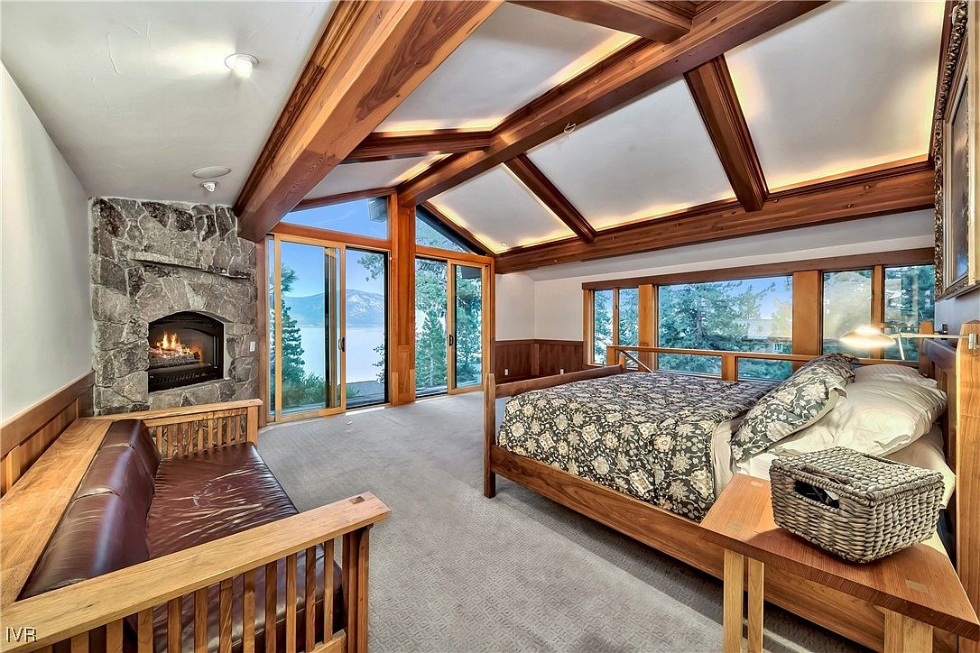 This primary bedroom features a stone fireplace, a seating area with a leather couch, and a large bed with a patterned comforter. The room has a vaulted ceiling with wooden beams and large windows offering a view of the lake and trees. The overall style is rustic and luxurious.