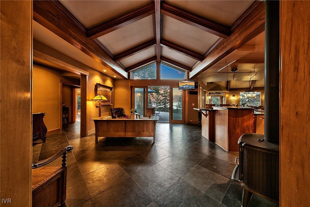 This interior shot showcases a spacious living room with a vaulted ceiling featuring exposed wooden beams. The room is furnished with a large wooden desk, and a stone tile floor adds a rustic touch. Sliding glass doors lead to an outdoor area, providing natural light and a view.
