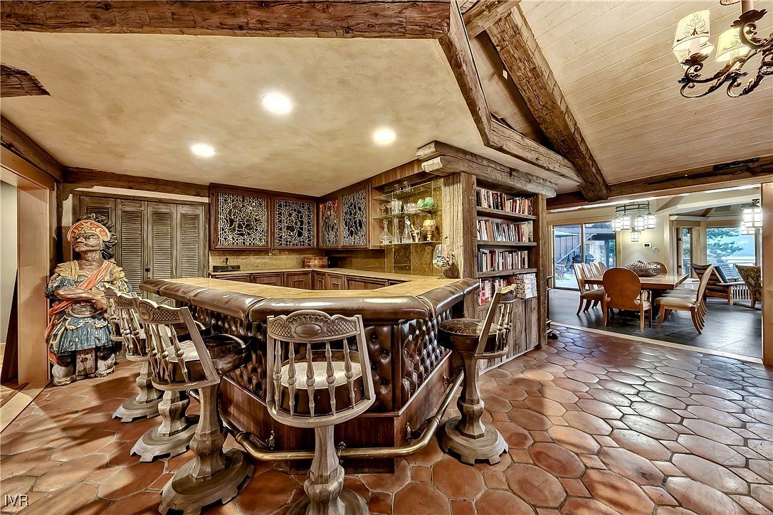 This image showcases a unique kitchen with a rustic and vintage aesthetic. The focal point is a custom-built bar with leather upholstery and ornate wooden chairs. The kitchen features wooden cabinetry with decorative glass panels, exposed wooden beams, and a distinctive stone tile floor, creating a warm and inviting atmosphere.