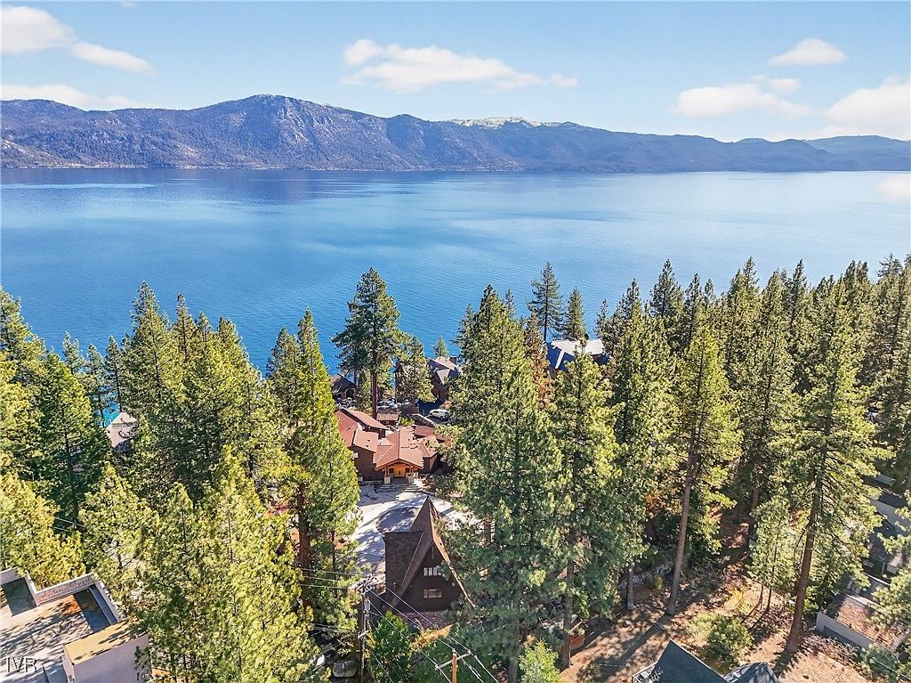 This aerial shot showcases a collection of houses nestled among tall evergreen trees, with a stunning view of a large, calm lake and distant mountains. The houses feature varying architectural styles, with some having visible rooflines and others partially obscured by the dense foliage. The overall impression is one of serene lakeside living in a natural setting.
