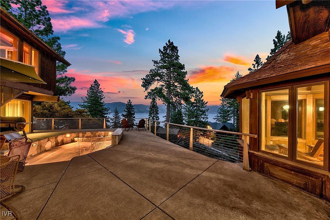 This image showcases a spacious patio or deck area of a luxurious home at sunset. The area features a stone fire pit, comfortable seating, and a modern railing system, all overlooking a scenic view of a lake and mountains. The warm lighting and beautiful sky create an inviting and upscale atmosphere, perfect for outdoor entertaining.