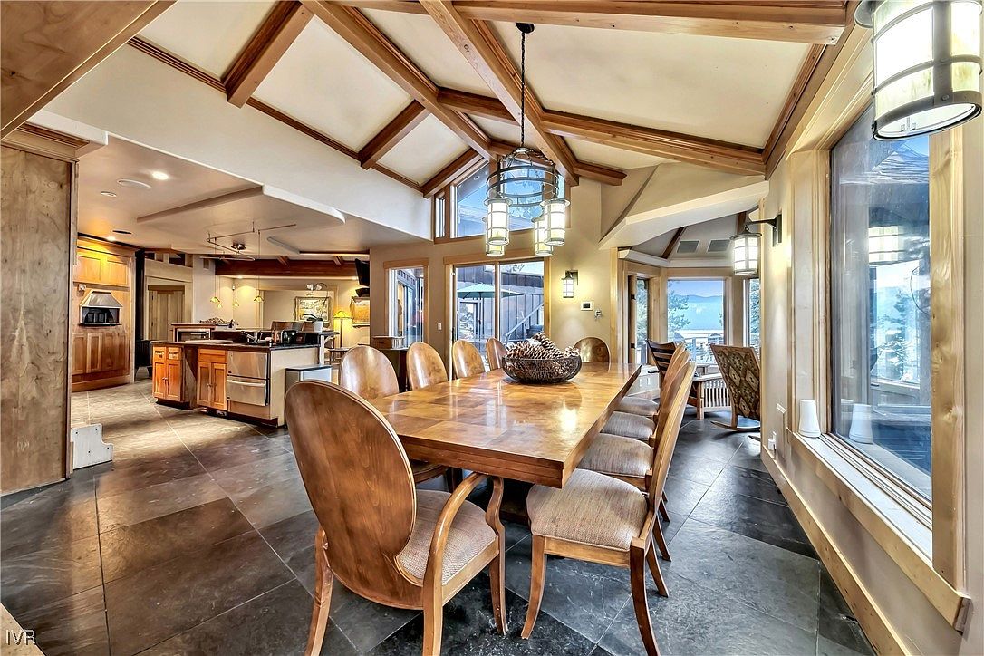 This is an interior shot of a dining room featuring a large wooden dining table with upholstered chairs. The room has a high ceiling with exposed wooden beams and a decorative chandelier. Large windows provide natural light and views of the outdoors, while the flooring is a dark, patterned tile. The style is rustic and elegant.