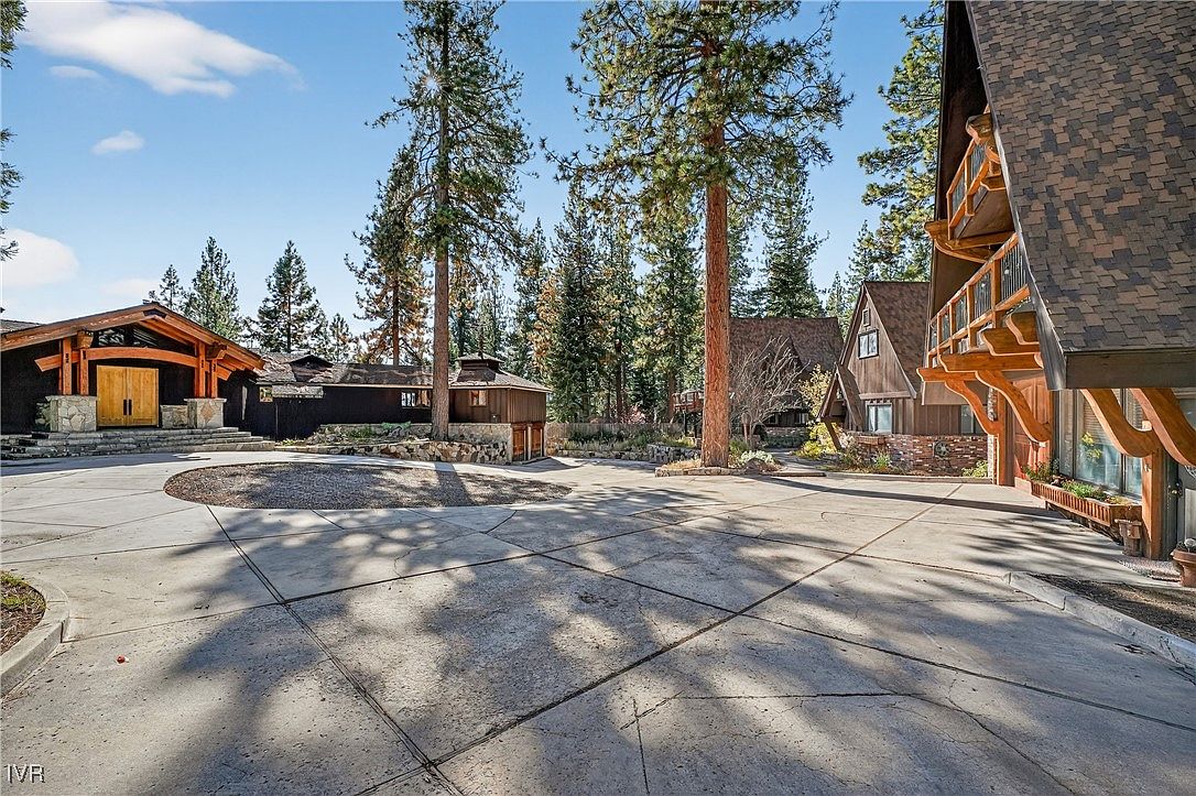 This is a front view of a luxurious mountain home with a large concrete driveway. The property features multiple structures with a mix of architectural styles, including A-frame elements and rustic wood finishes. Mature trees surround the buildings, creating a sense of privacy and seclusion.