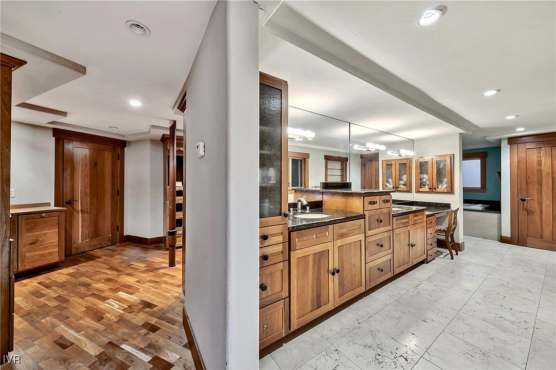 This is a well-appointed primary bathroom featuring wood cabinetry with granite countertops and a large mirror. The flooring is marble tile, and there are two wooden doors, one leading to a closet or dressing area. The overall impression is luxurious and spacious.