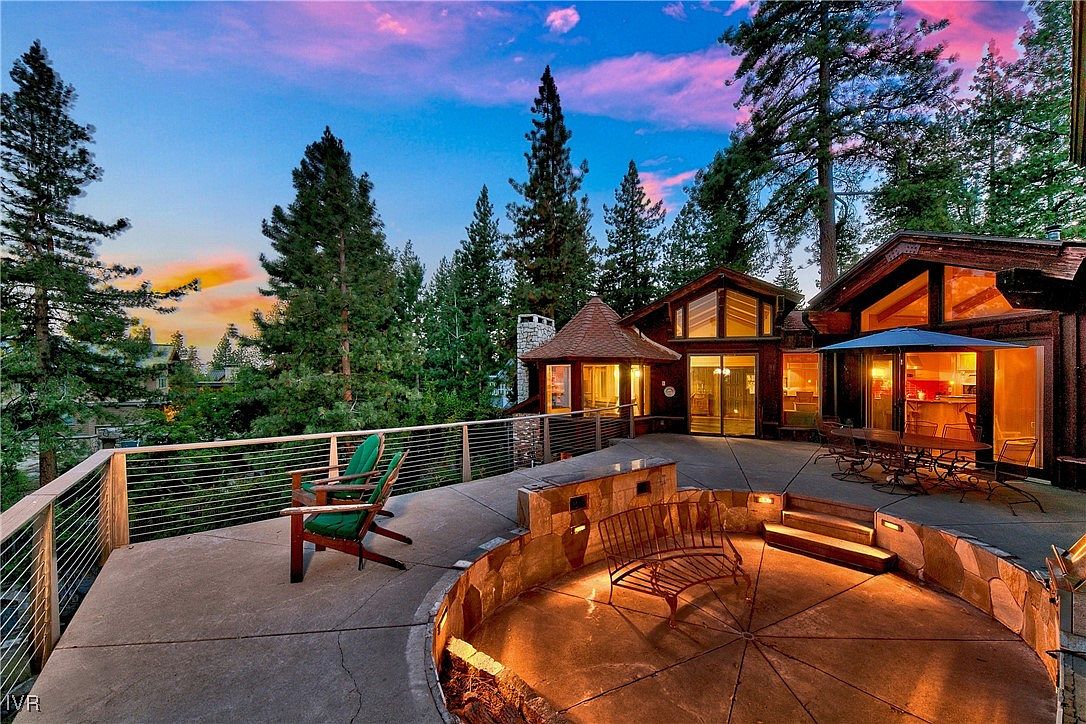 This image showcases a luxurious outdoor living space featuring a spacious patio with a unique sunken fire pit area surrounded by stone seating. The patio is furnished with comfortable seating and a dining set, perfect for entertaining. The backdrop includes a beautifully designed home with large windows and lush greenery, creating a serene and inviting atmosphere.
