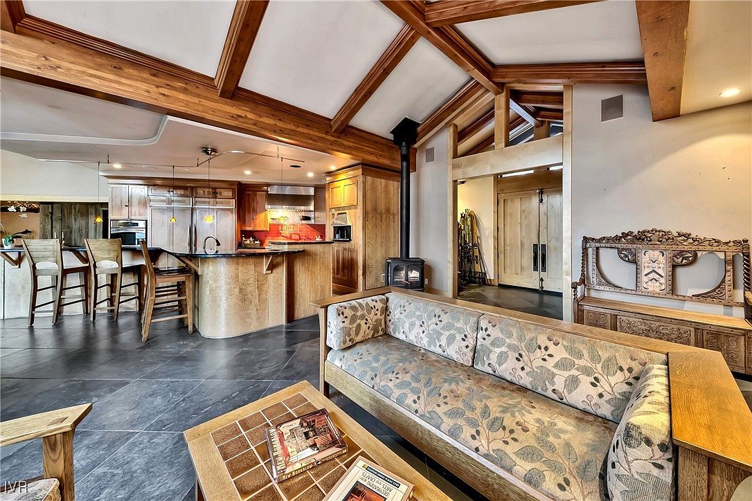 This interior shot showcases a cozy living room that seamlessly blends into an open-concept kitchen. The living area features a comfortable sofa with a floral pattern, a wooden coffee table, and a striking wood-burning stove. The kitchen boasts wooden cabinetry, a curved island, and bar seating, all under a ceiling with exposed wooden beams, creating a warm and inviting atmosphere.