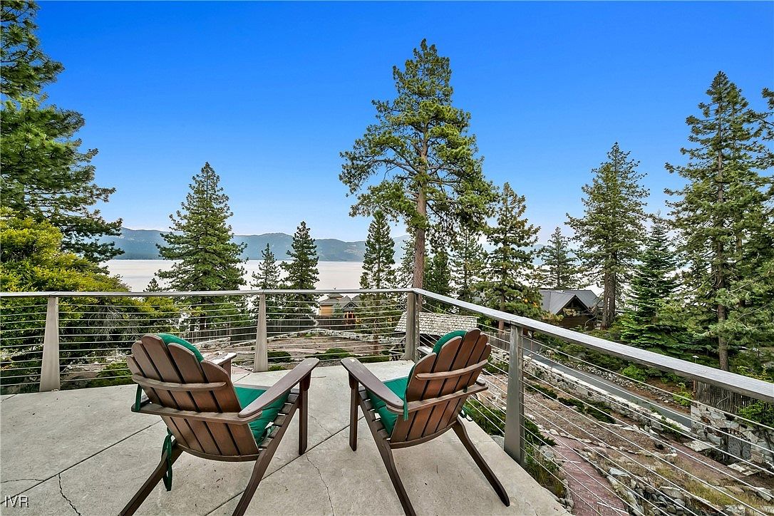This image showcases a serene outdoor patio or deck area, featuring two wooden Adirondack chairs with green cushions, inviting relaxation with a view. The deck is surrounded by a modern railing system with cable wires, offering unobstructed views of the lake and surrounding trees. The setting suggests a peaceful retreat, perfect for enjoying the natural beauty of the location.