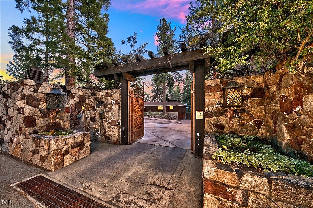 This image showcases an impressive entryway featuring a stone wall and a wooden pergola-style gate. The gate opens to reveal a glimpse of a house in the background, framed by trees. The stone and wood elements create a rustic and inviting atmosphere, suggesting a property with character and charm.