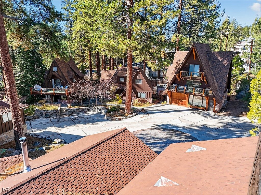 This image showcases the front view of several A-frame style cabins nestled among tall pine trees. A concrete driveway provides ample parking space, leading up to the garages and entrances of the cabins. The architecture blends rustic charm with modern design, creating an inviting mountain retreat.