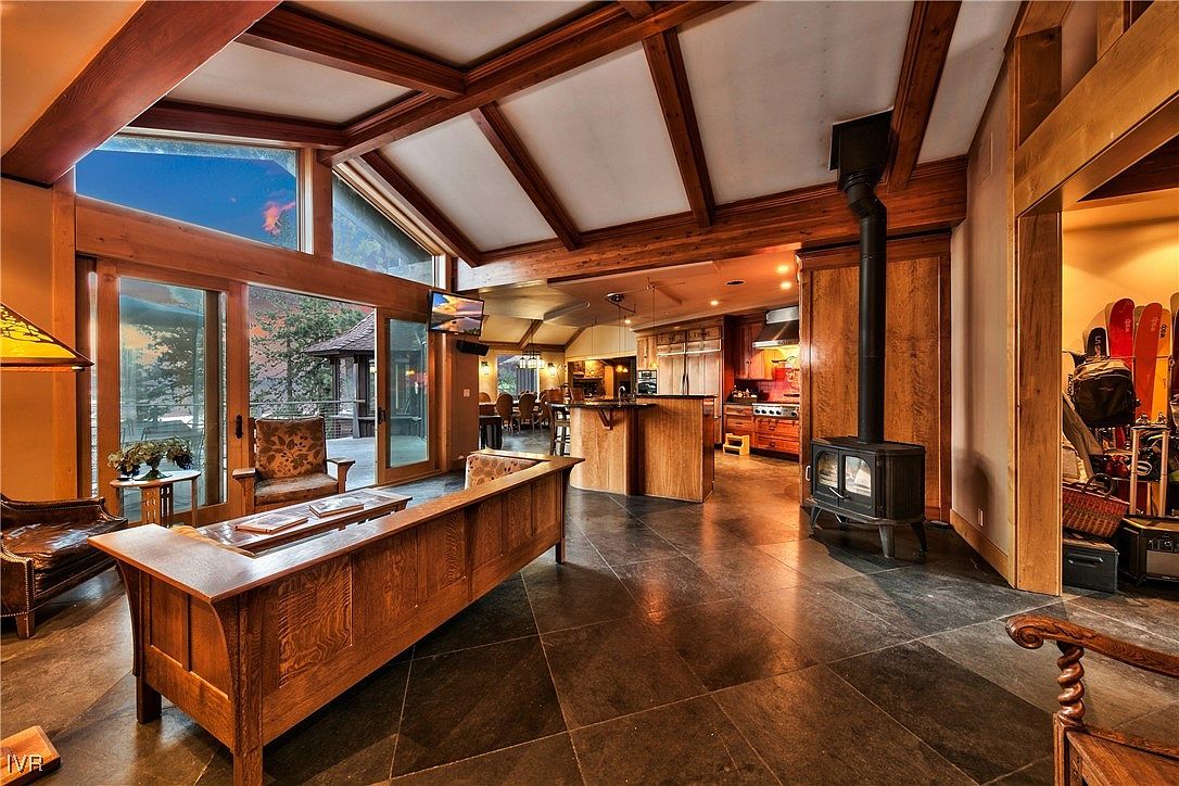 This interior shot showcases a spacious living area with a rustic yet elegant design. The room features a stone tile floor, exposed wooden beams, and large windows offering natural light and views. A wood-burning stove adds a cozy touch, while the open layout seamlessly connects to the kitchen and dining area, creating an inviting space for relaxation and entertainment.