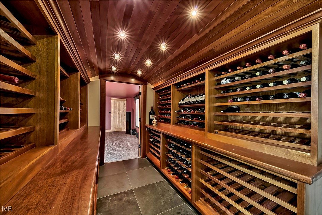This is an interior shot of a well-stocked wine cellar, featuring custom wooden shelving filled with numerous bottles of wine. The ceiling is also wood-paneled and has recessed lighting, creating a warm and inviting atmosphere. The floor is tiled, and an open doorway leads to another room, suggesting ample space and a sophisticated design.