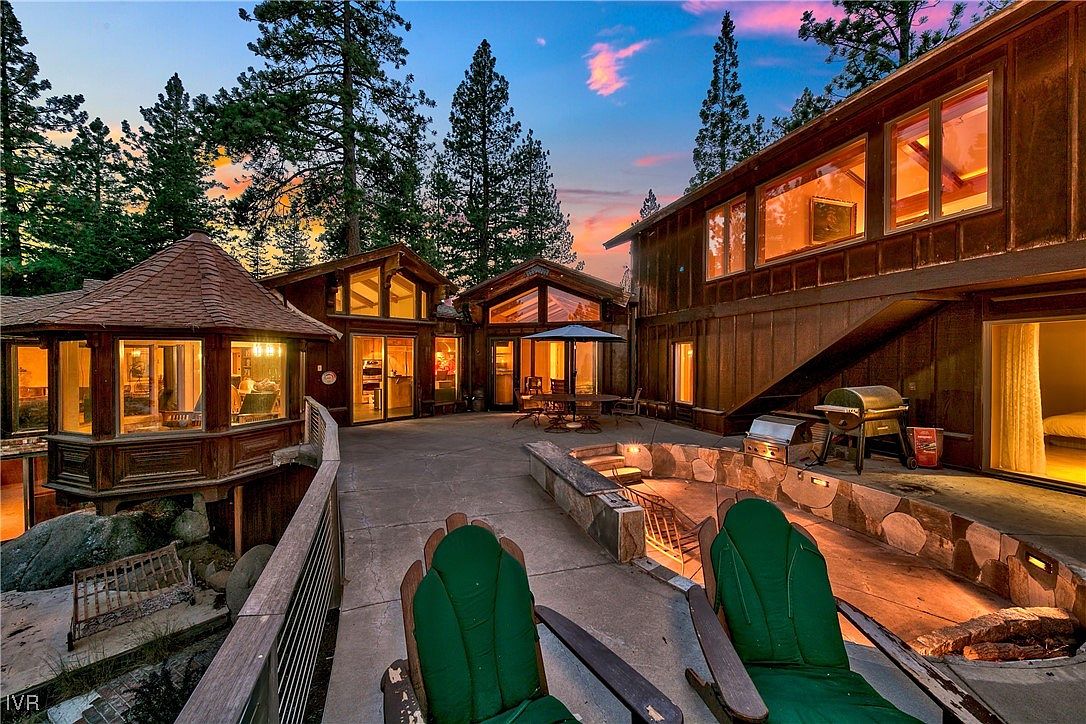 This image showcases a spacious patio area of a luxurious mountain home at dusk. The patio features comfortable outdoor seating, a built-in fire pit, and a grilling area, perfect for entertaining. The warm lighting from the home's windows creates an inviting atmosphere, highlighting the property's appeal for relaxation and outdoor living.