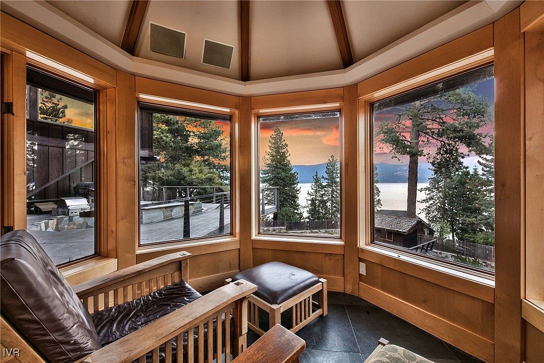 This sunroom features a cozy seating area with a leather chair and ottoman, surrounded by large windows offering panoramic views of the lake and surrounding landscape. The room is finished with wood paneling and a unique ceiling design, creating a warm and inviting atmosphere. The dark tile flooring adds a touch of elegance to the space.