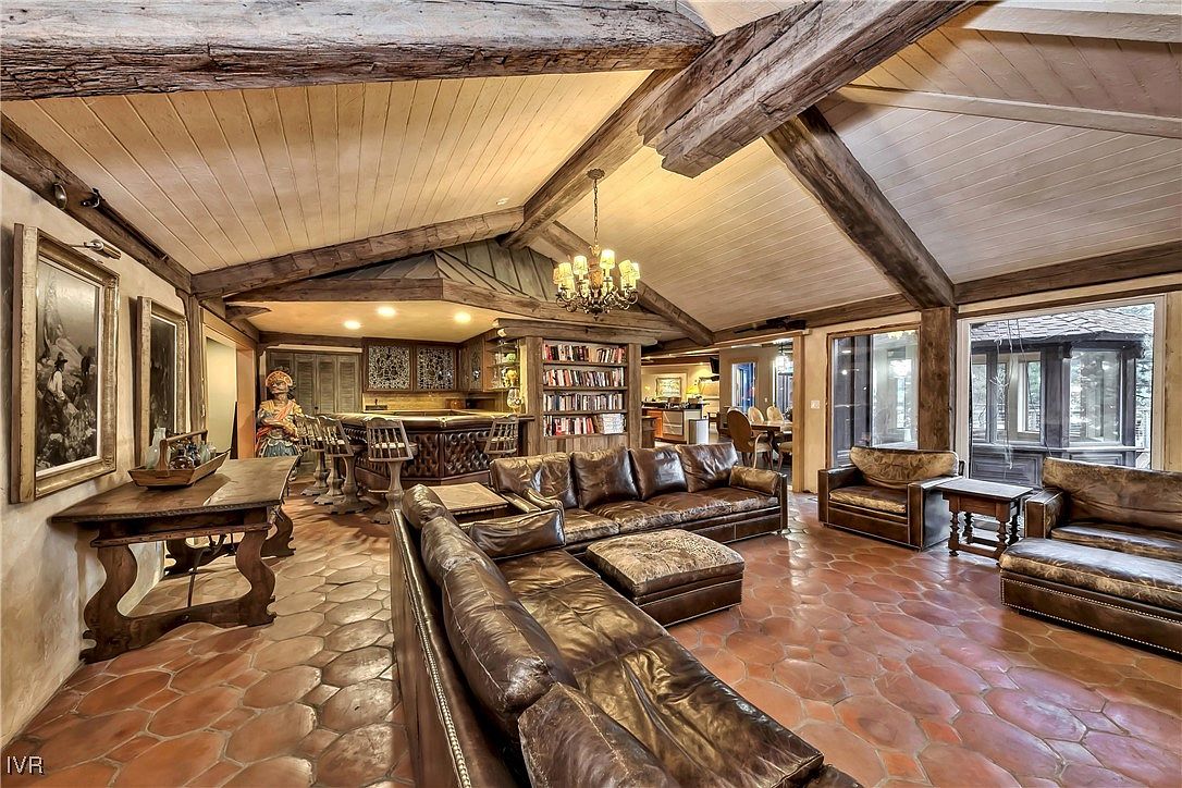 This is an interior shot of a rustic living room featuring exposed wooden beams, a large leather sectional sofa, and terracotta tile flooring. The room includes a bar area with ornate wood detailing and a bookshelf, suggesting a cozy and luxurious space for entertaining. The perspective is from the front of the room, showcasing the depth and layout.