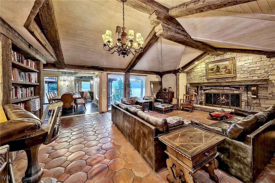 This is a warm and inviting living room featuring exposed wooden beams, a stone fireplace, and comfortable leather furniture. The room is well-lit by a chandelier and natural light from the windows, which offer a glimpse of the outdoors. The overall style is rustic and luxurious, creating a cozy and sophisticated atmosphere.