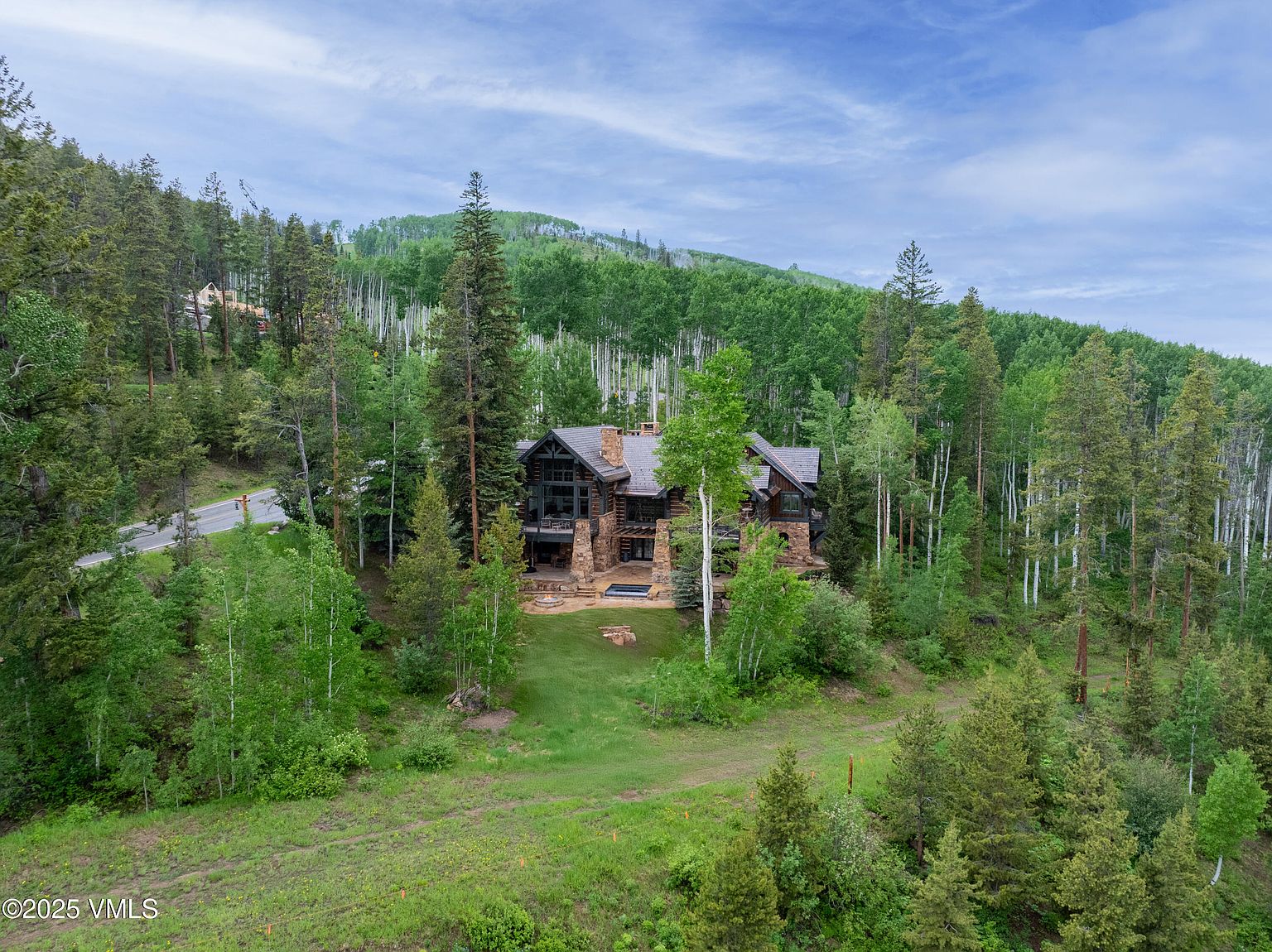 This aerial view showcases a stunning log cabin nestled amidst a lush forest. The home features a stone chimney, a dark roof, and a well-maintained lawn with a patio area. The surrounding landscape includes a mix of evergreen and deciduous trees, creating a serene and private setting.