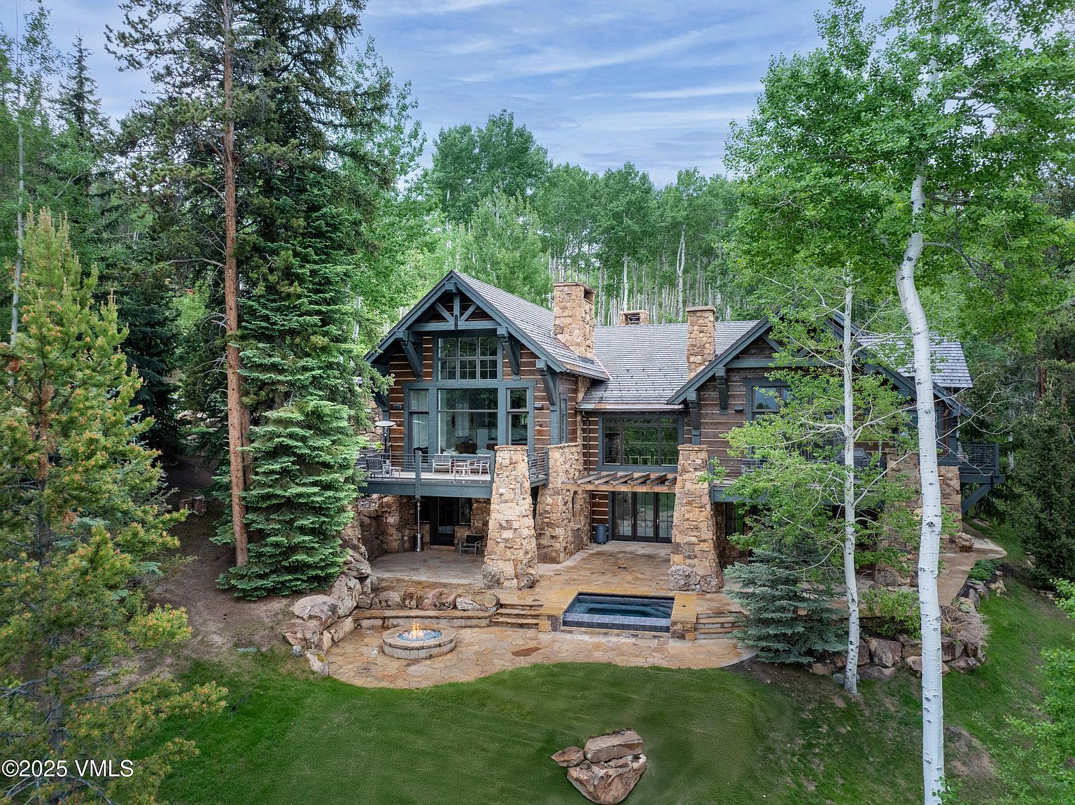 This is a rear exterior view of a luxurious log cabin-style home nestled among lush trees. The house features stone pillars supporting a deck, a stone patio with a fire pit and hot tub, and large windows offering views of the surrounding landscape. The overall impression is one of rustic elegance and secluded comfort.