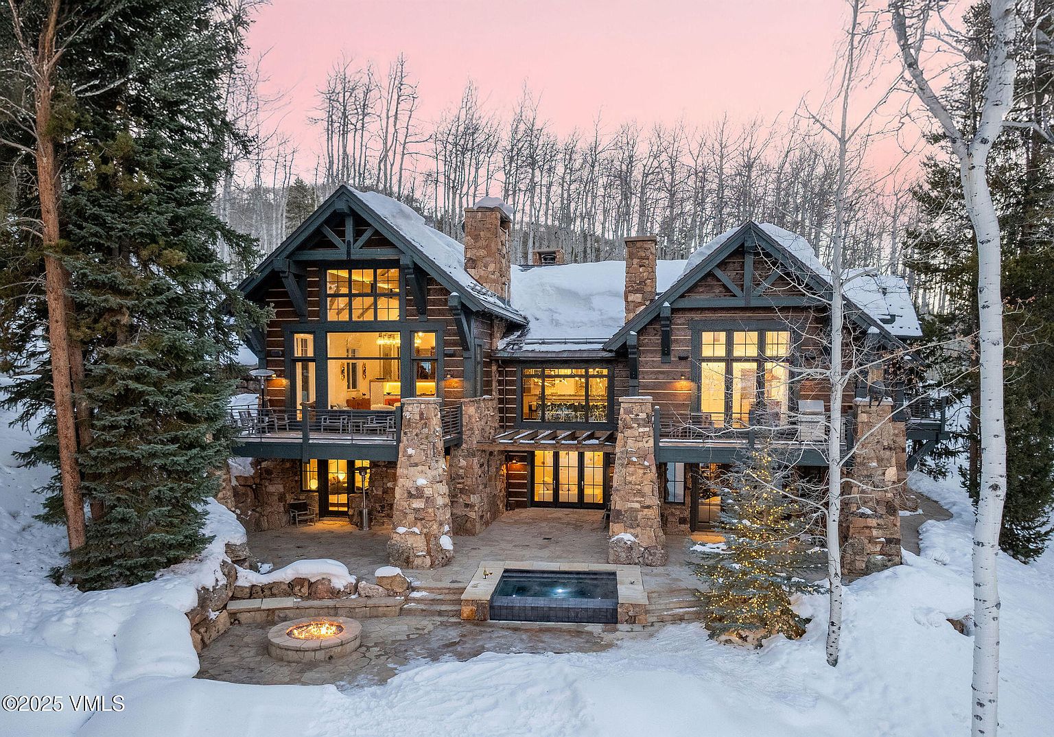 This is a stunning rear view of a luxurious mountain home, showcasing its grand architecture and outdoor amenities. The home features a stone foundation, log siding, large windows, and multiple balconies. A patio area includes a hot tub and a fire pit, perfect for enjoying the snowy landscape and creating a cozy atmosphere.