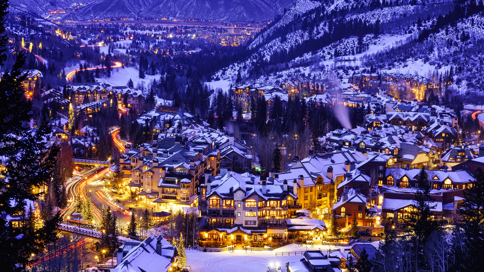 This aerial view showcases a luxurious ski resort town at night, blanketed in snow. The warm glow of lights emanating from homes and businesses creates a cozy and inviting atmosphere, contrasting with the cool blue tones of the surrounding mountains and the twinkling lights of a distant town. A winding road with light trails adds dynamic movement to the scene.