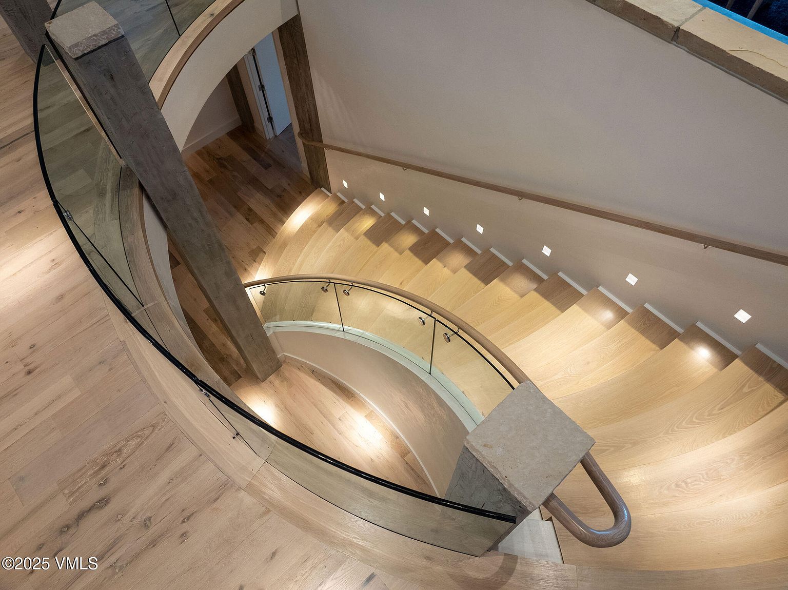 This is a high-angle shot of a modern, curved staircase. The stairs are made of light-colored wood and feature integrated lighting along the wall. A glass railing with black trim follows the curve of the staircase, and a wooden handrail is mounted on the wall, creating a sleek and contemporary design.