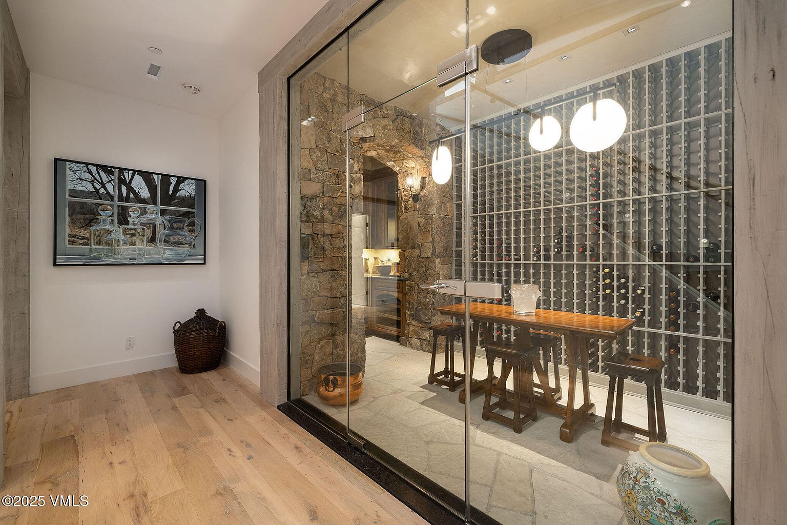 This image showcases a sophisticated wine cellar, featuring a stone wall accent, a glass-enclosed space, and a wooden tasting table with stools. The wine racks are filled with bottles, illuminated by pendant lights, creating an inviting atmosphere for wine enthusiasts. The flooring appears to be stone tile, adding to the cellar's rustic yet elegant charm.