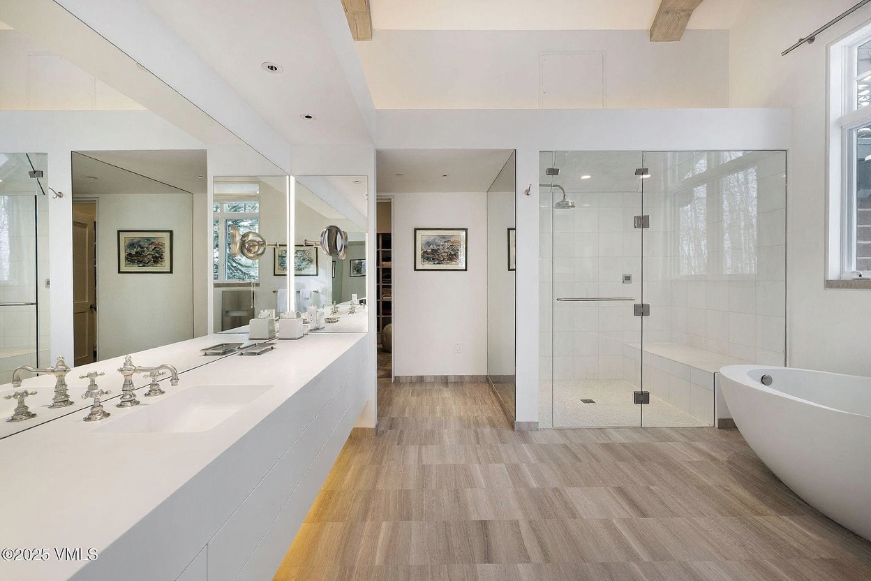 This is a well-lit primary bathroom featuring a long, sleek white countertop with dual sinks and modern fixtures. A large, glass-enclosed shower with a built-in bench complements the freestanding bathtub, creating a spa-like atmosphere. The neutral-toned tile flooring adds a touch of elegance to the space.