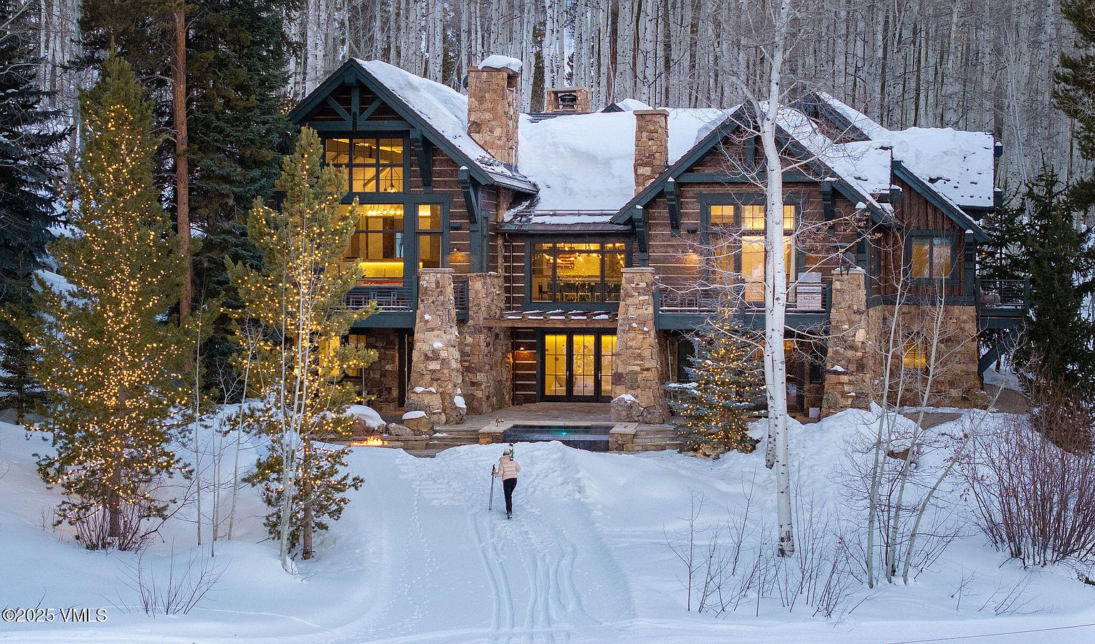 This is a front exterior view of a luxurious log cabin home in a snowy, wooded setting. The home features stone accents, multiple gabled roofs covered in snow, and large windows that suggest a spacious interior. A person is seen cross-country skiing towards the house, adding a sense of scale and activity.