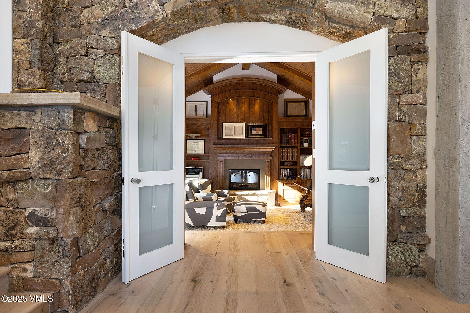 This image showcases an inviting office or study, viewed through a wide doorway with white framed glass doors. The room features a stone archway, hardwood floors, a fireplace with a wooden mantel, and built-in bookshelves. The decor includes a patterned armchair and ottoman, creating a cozy and sophisticated atmosphere.