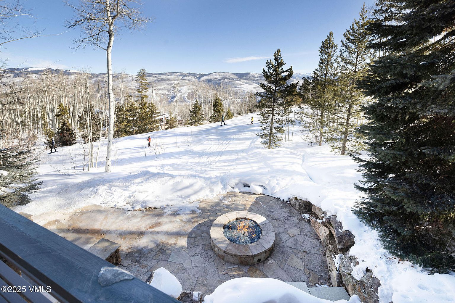 This image showcases an inviting outdoor patio area with a stone fire pit as its focal point, surrounded by a stone-paved surface. The patio overlooks a snow-covered landscape with scattered trees and distant mountains, creating a serene and picturesque setting. The view is from an elevated position, offering a wide perspective of the outdoor space and the natural beauty beyond.