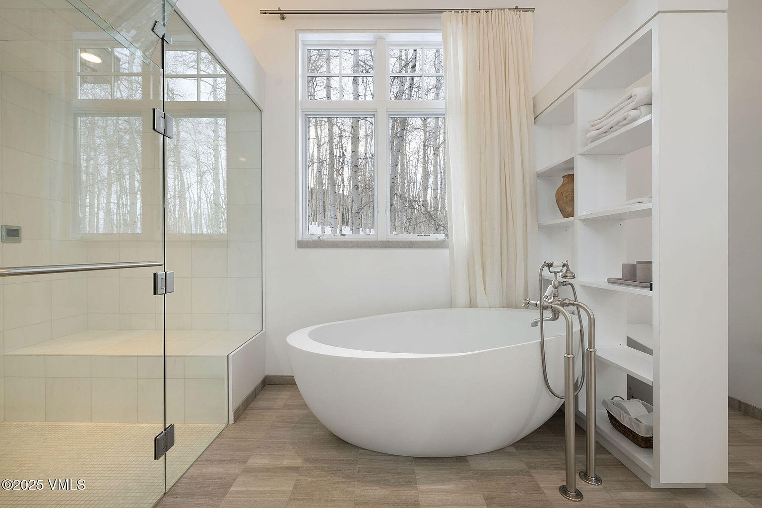 This is a bright and modern primary bathroom featuring a freestanding white bathtub positioned near a large window with a view of a birch tree forest. A glass-enclosed shower with a built-in bench is on the left, and a built-in shelving unit provides storage on the right. The flooring is a light-toned tile, and the overall aesthetic is clean, minimalist, and spa-like.